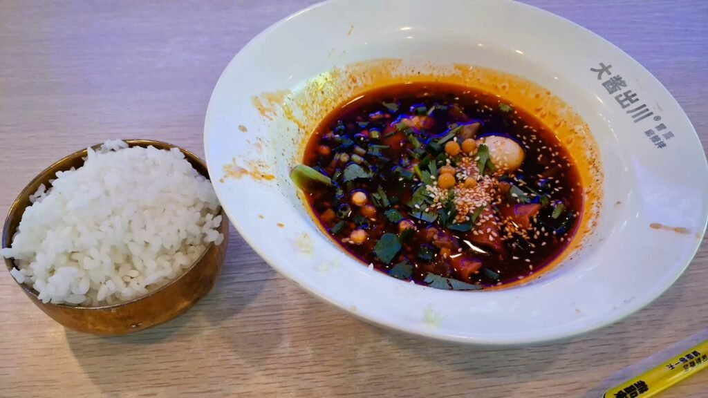A bowl of malatang spicy numbing hot pot with dark red broth, mixed ingredients and sesame seeds, served with steamed rice in Hainan China