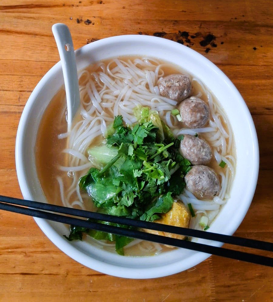 Top-down view of a bowl of Hainan rice noodle soup with beef meatballs, greens and coriander