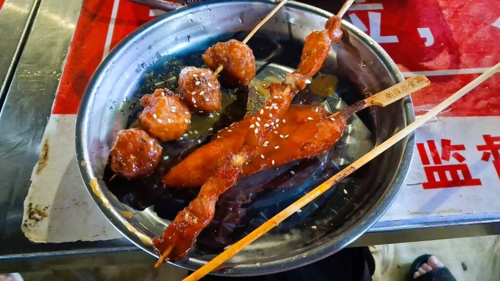 Glazed meat and meatball skewers with sesame seeds in a metal bowl at a Chinese night market barbecue stall in Hainan