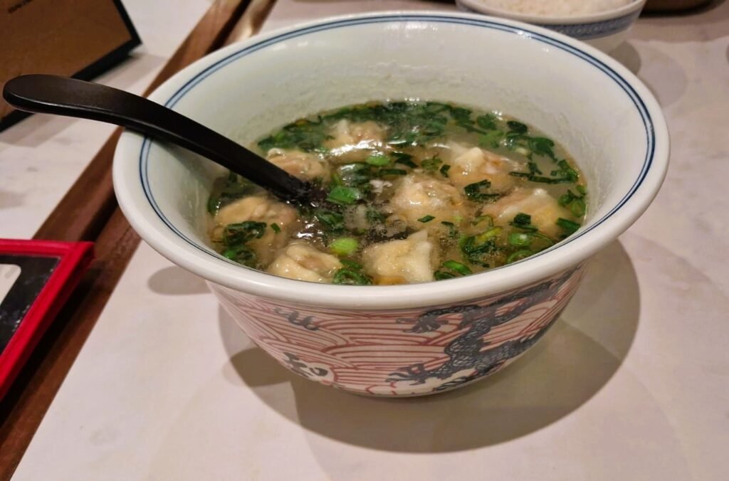A ceramic bowl of wonton soup with green herbs and clear broth at a Chinese restaurant in Hainan